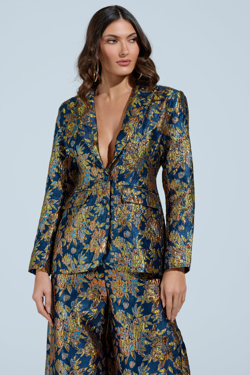 A person in AKIRA's SURRENDER FLORAL BLAZER, featuring a deep blue floral pattern with gold and green accents, stands against a light backdrop. Their hair is styled in loose waves as they glance to the side.