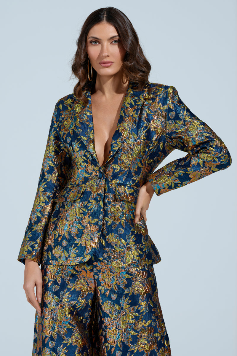 A person with long, wavy hair poses confidently in the SURRENDER FLORAL BLAZER by AKIRA Label, featuring intricate gold and green patterns. The metallic material shimmers subtly against a plain light blue backdrop, highlighting the blazer's vibrant design.