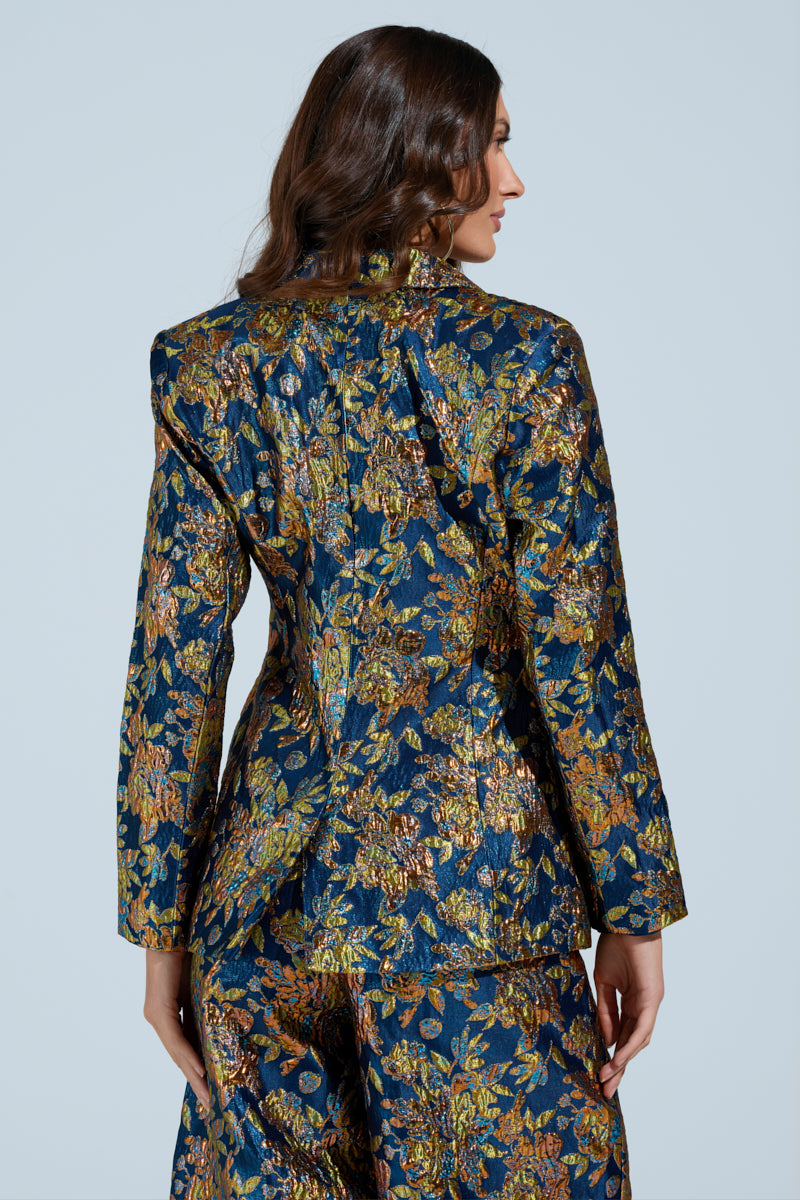 A person with long brown hair is wearing the SURRENDER FLORAL BLAZER from AKIRA Label, adorned with an ornate metallic floral pattern, paired with pants. They stand against a light gray background, facing away from the camera.