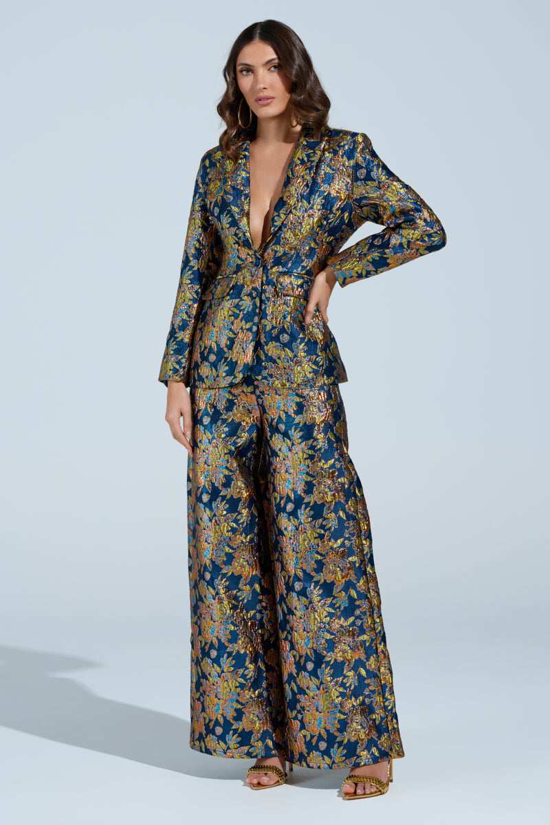 A woman confidently poses in the AKIRA Label's SURRENDER FLORAL BLAZER pantsuit, showcasing a luxurious dark blue brocade with intricate floral patterns and metallic accents. The deep V-neck blazer is paired with wide-leg trousers against a light, neutral background.