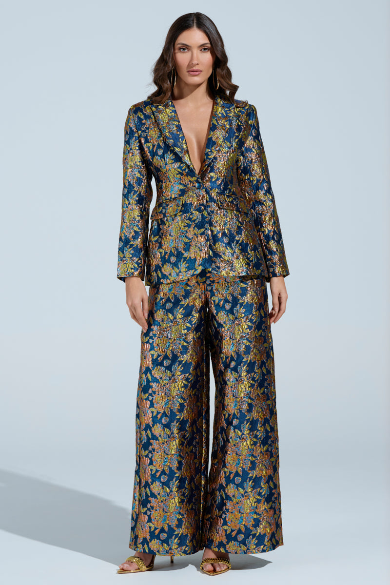 A woman poses against a light blue background wearing the AKIRA Label SURRENDER FLORAL BLAZER with a matching pantsuit. The shiny blazer and deep V-neck wide-leg trousers feature metallic hues of blue, gold, and pink. Her long, wavy hair complements her open-toed heels.