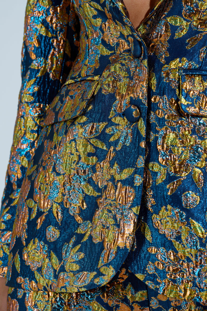 A close-up of the SURRENDER FLORAL BLAZER reveals a richly textured brocade pattern in vibrant blue, green, and gold, with detailed embroidery featuring shimmering metallic threads that create an intricate and luxurious design.
