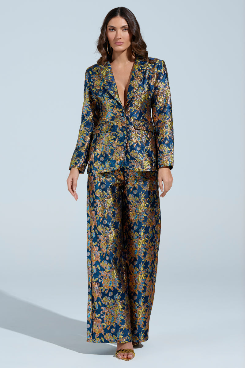 Wearing the SURRENDER FLORAL BLAZER, a person exudes confidence in an ornate, floral-patterned dark blue pantsuit with gold and bronze metallic details. With long, wavy hair and open-toe shoes, they stand against a plain light blue background.