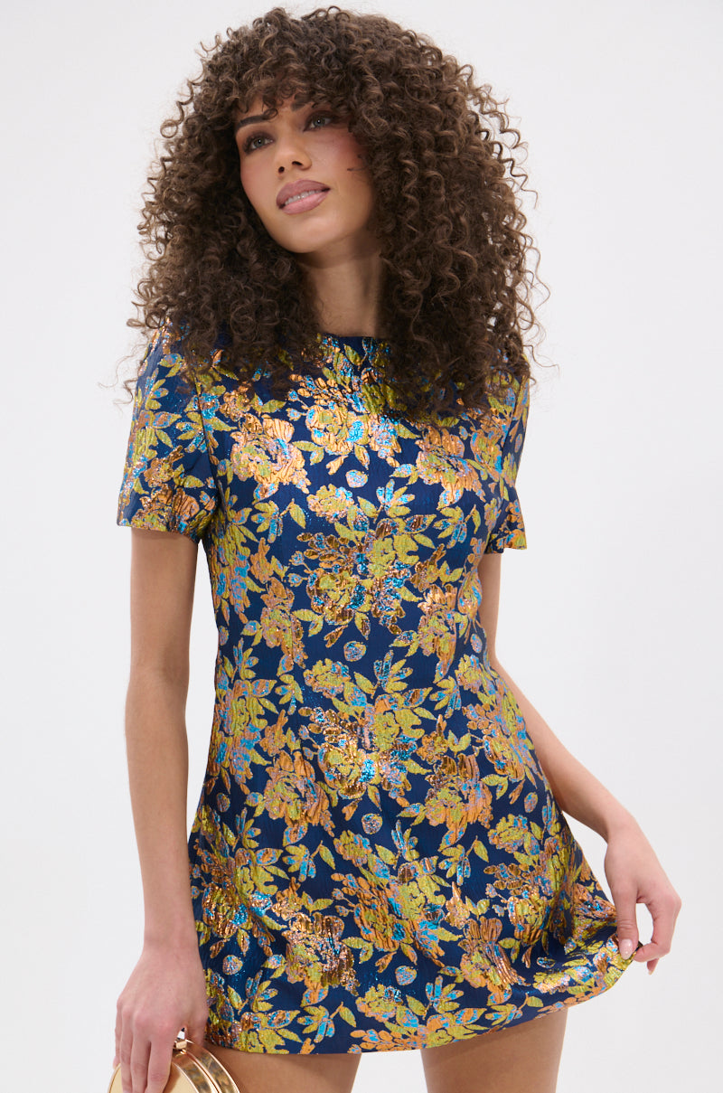 A woman with curly hair wears the BLUE IS THE NEW BLACK BROCADE MINI DRESS, a navy metallic mini dress with a shiny gold and orange floral pattern, and holds a small round gold handbag against a plain white background.