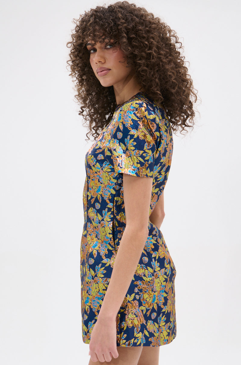 A woman with curly hair wears the BLUE IS THE NEW BLACK BROCADE MINI DRESS. The short-sleeved dress features a colorful floral pattern on a dark background. She stands sideways, looking at the camera, against a plain white backdrop.