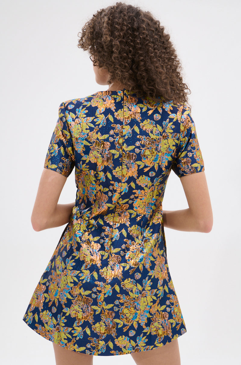 A woman with curly hair models the BLUE IS THE NEW BLACK BROCADE MINI DRESS, a short-sleeved blue mini dress with a gold, orange, and green floral metallic brocade pattern. She is turned away from the camera, against a plain white background.