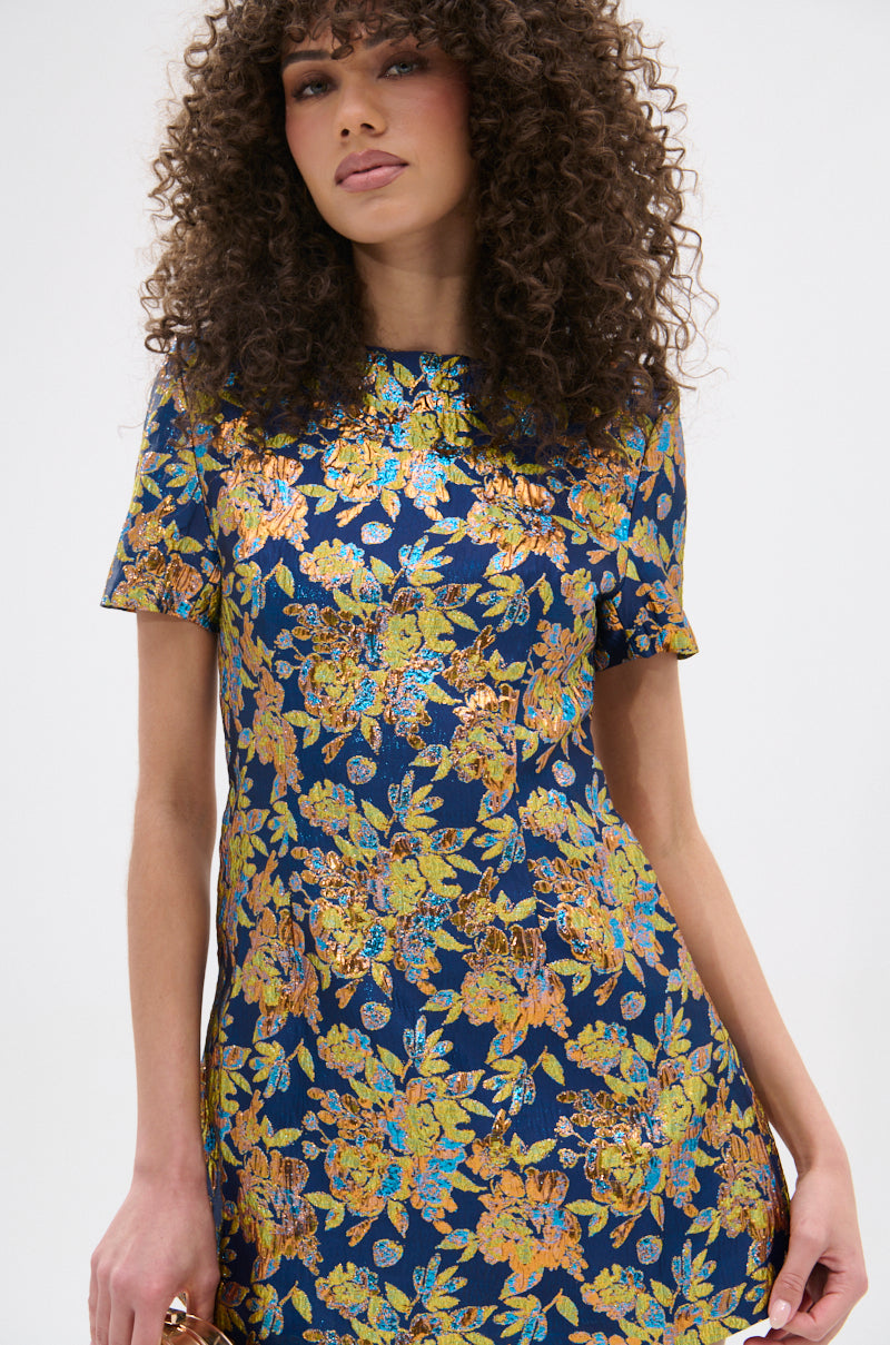 A woman with curly hair models the BLUE IS THE NEW BLACK BROCADE MINI DRESS, a navy blue skater dress with intricate gold and yellow floral patterns, standing against a plain white background.
