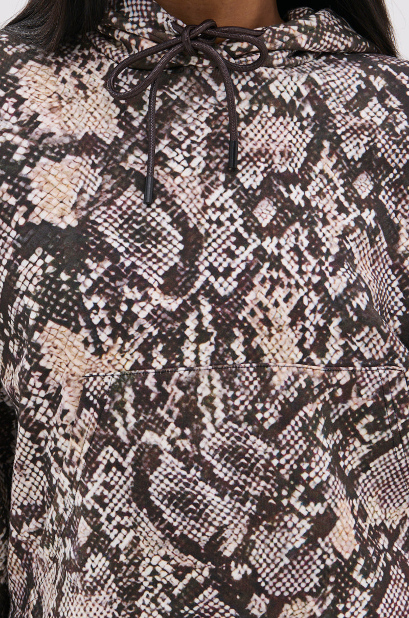 A close-up shows a person wearing the oversized PYTHON HOODED SWEATSHIRT, featuring a brown, black, and beige snake print. The hood’s drawstrings are visible near the top. 9