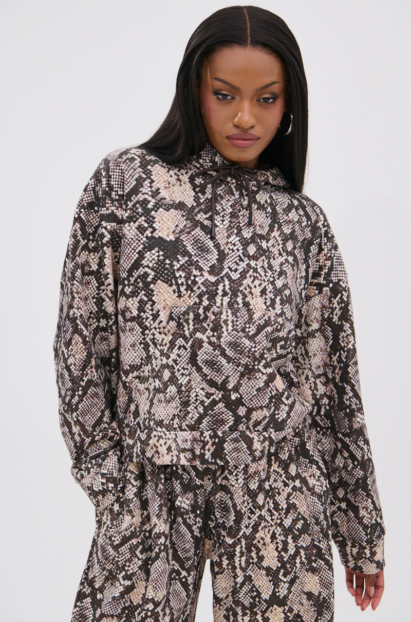 A woman models the PYTHON HOODED SWEATSHIRT with a brown and beige snakeskin pattern, standing against a white background and looking slightly downward with one hand in her pocket. 1