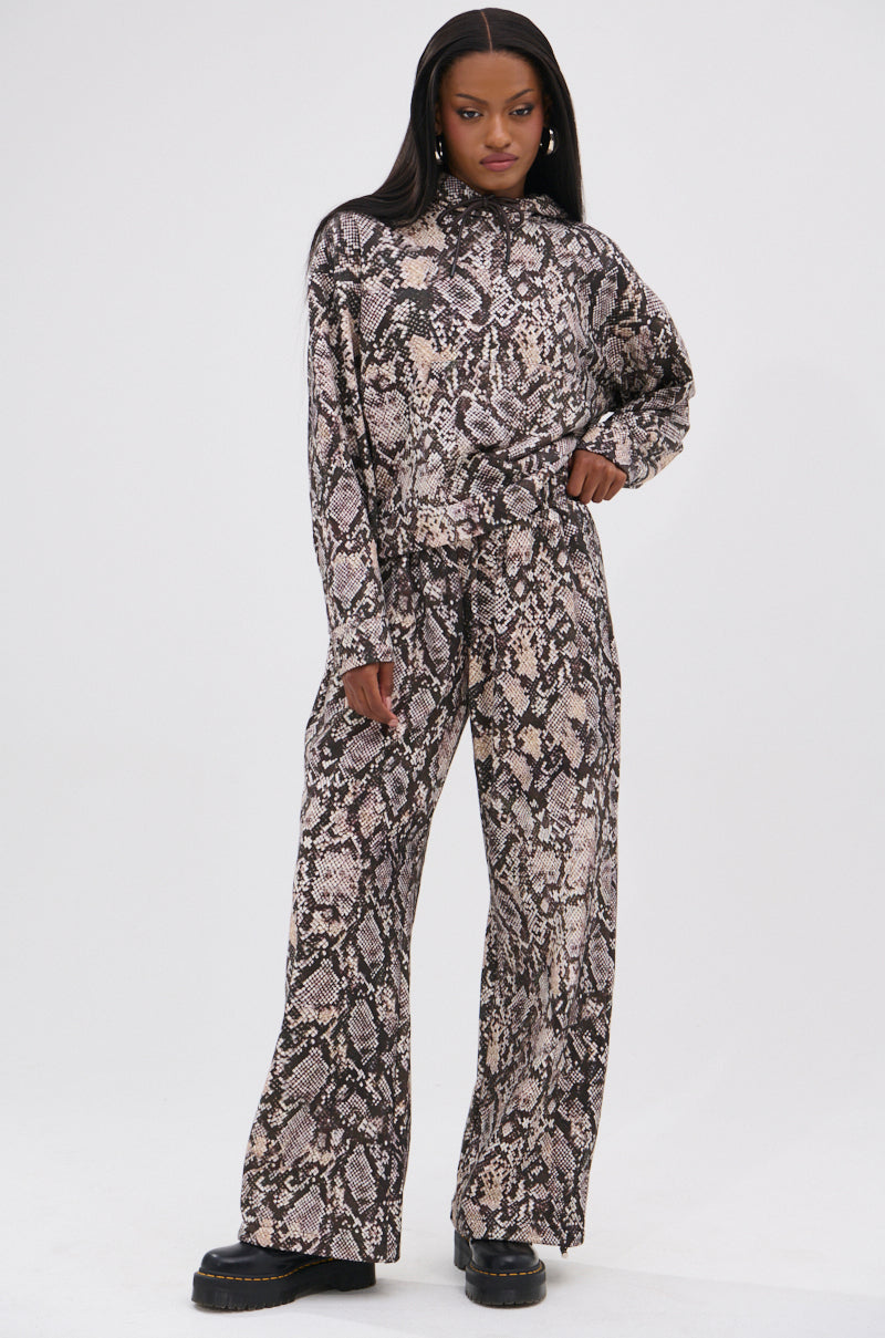 A woman confidently wears the Python Wide Leg Jogger, featuring an elastic waistband in gray and brown snake print, paired with a matching top and black platform shoes against a plain white background. 0