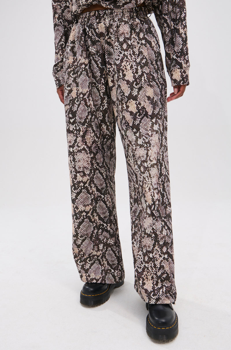 Model wears the PYTHON WIDE LEG JOGGER—loose-fitting, high-waisted pants with an elastic waistband in a brown and beige snake print—paired with a matching top and black platform shoes against a plain white background. 2