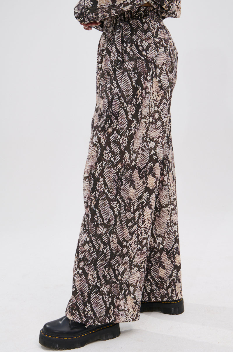 A person models the PYTHON WIDE LEG JOGGER featuring a brown and beige snake print and elastic waistband, paired with black chunky-soled shoes. Only the lower half is shown against a plain white background. 3
