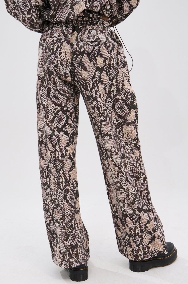 A model is shown from the back wearing the PYTHON WIDE LEG JOGGER—high-waisted, loose-fitting joggers with a brown and beige snake print and elastic waistband—paired with black chunky shoes, against a plain white background. 5