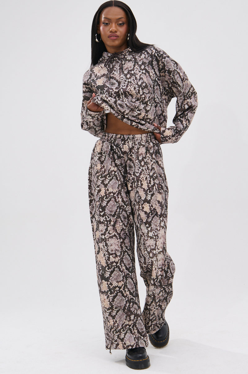 A woman poses confidently in the PYTHON WIDE LEG JOGGER, featuring an elastic waistband and brown-beige snake print, paired with a matching long-sleeve top and black platform shoes against a plain white background. 6