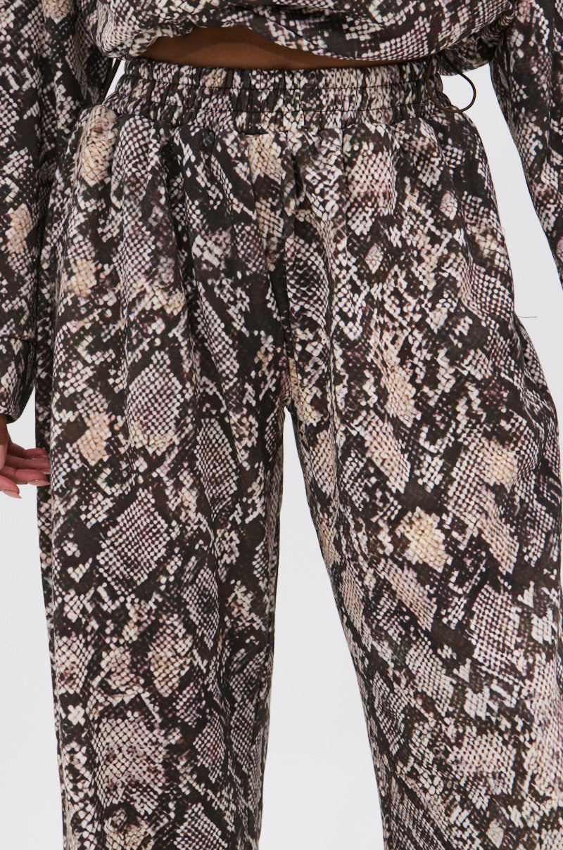 A close-up of a person wearing the PYTHON WIDE LEG JOGGER, featuring high-waisted, wide-leg pants with an elastic waistband in a brown, black, and beige snake print. Only the torso and upper legs are visible against a white background. 7