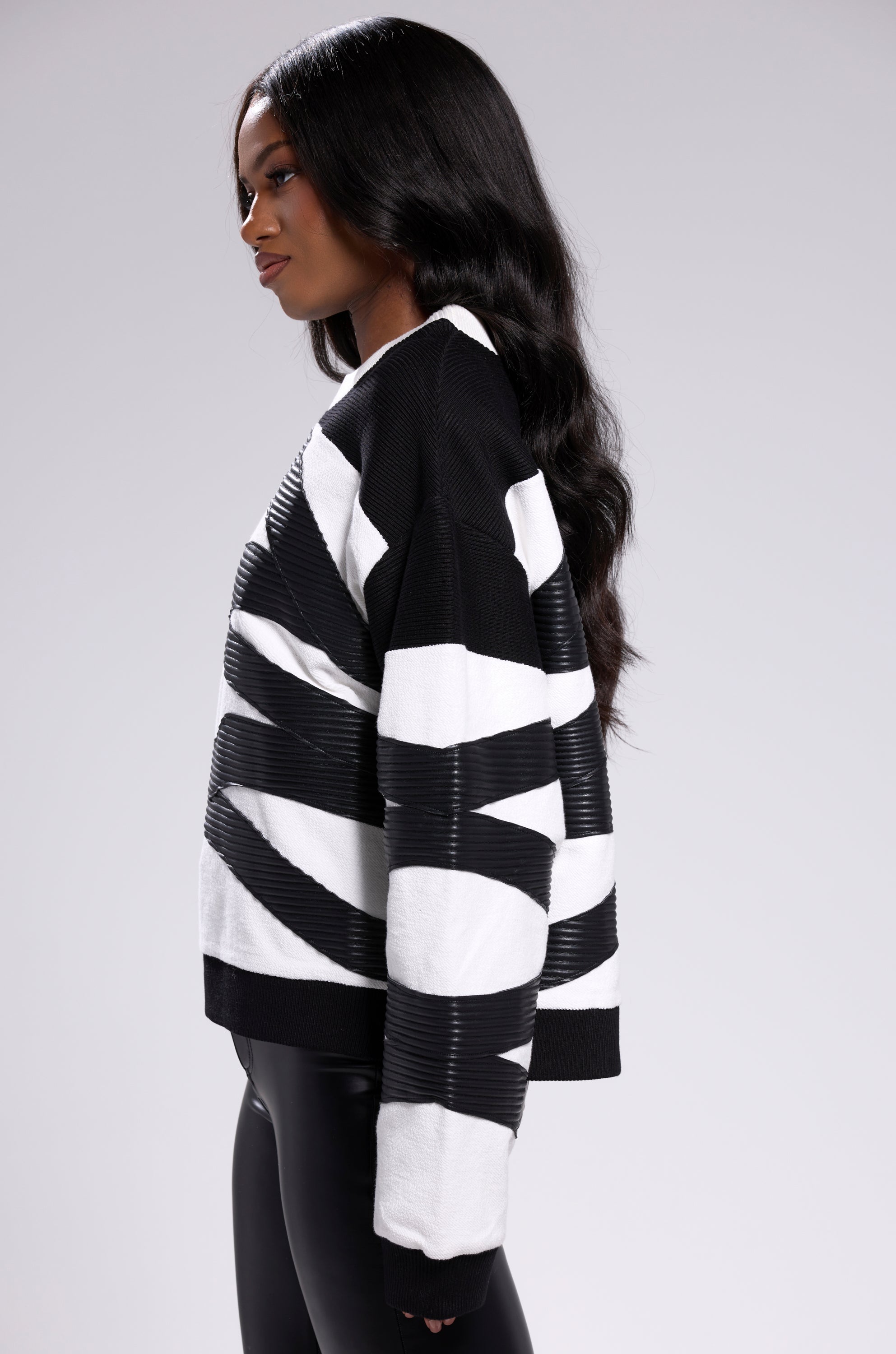 A woman with long wavy hair models the CROSSING LINES SWEATSHIRT with black leggings, standing in profile against a plain light gray background for a chic streetwear look.