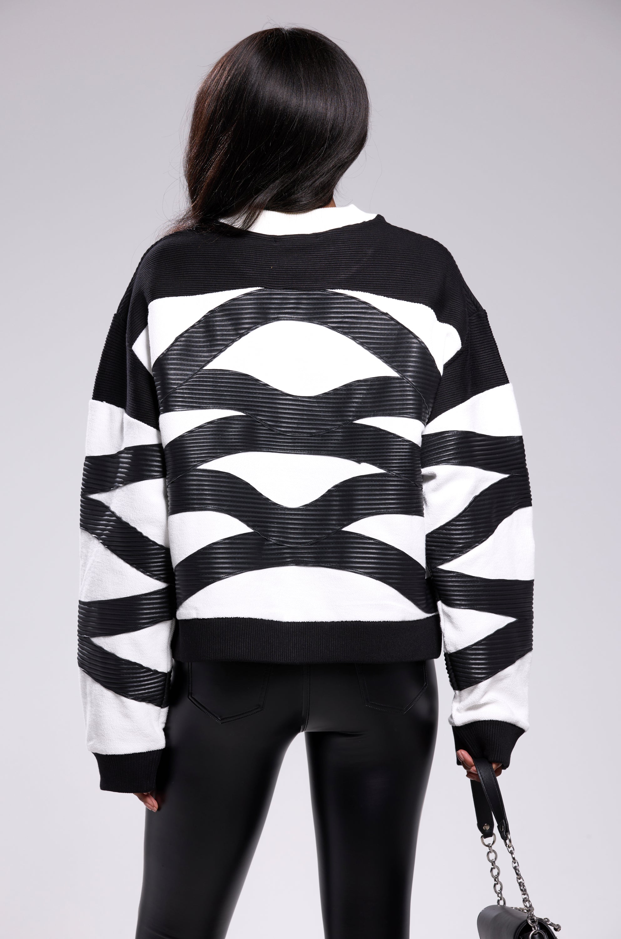 A person with long dark hair, seen from behind, wears the bold CROSSING LINES SWEATSHIRT with abstract colorblock patterns and shiny black pants, holding a small black purse—perfect for standout streetwear style.