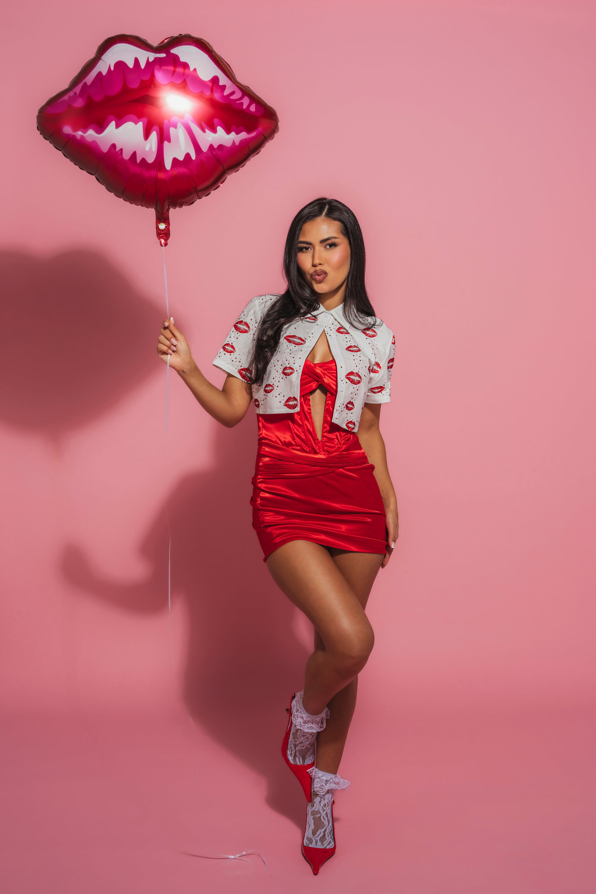 A woman models the KISS KISS RHINESTONE CROPPED BUTTON DOWN with a red lip-print dress, lace socks, and heels. She poses against a pink background holding a red lips-shaped balloon, one leg crossed in front of the other. 3