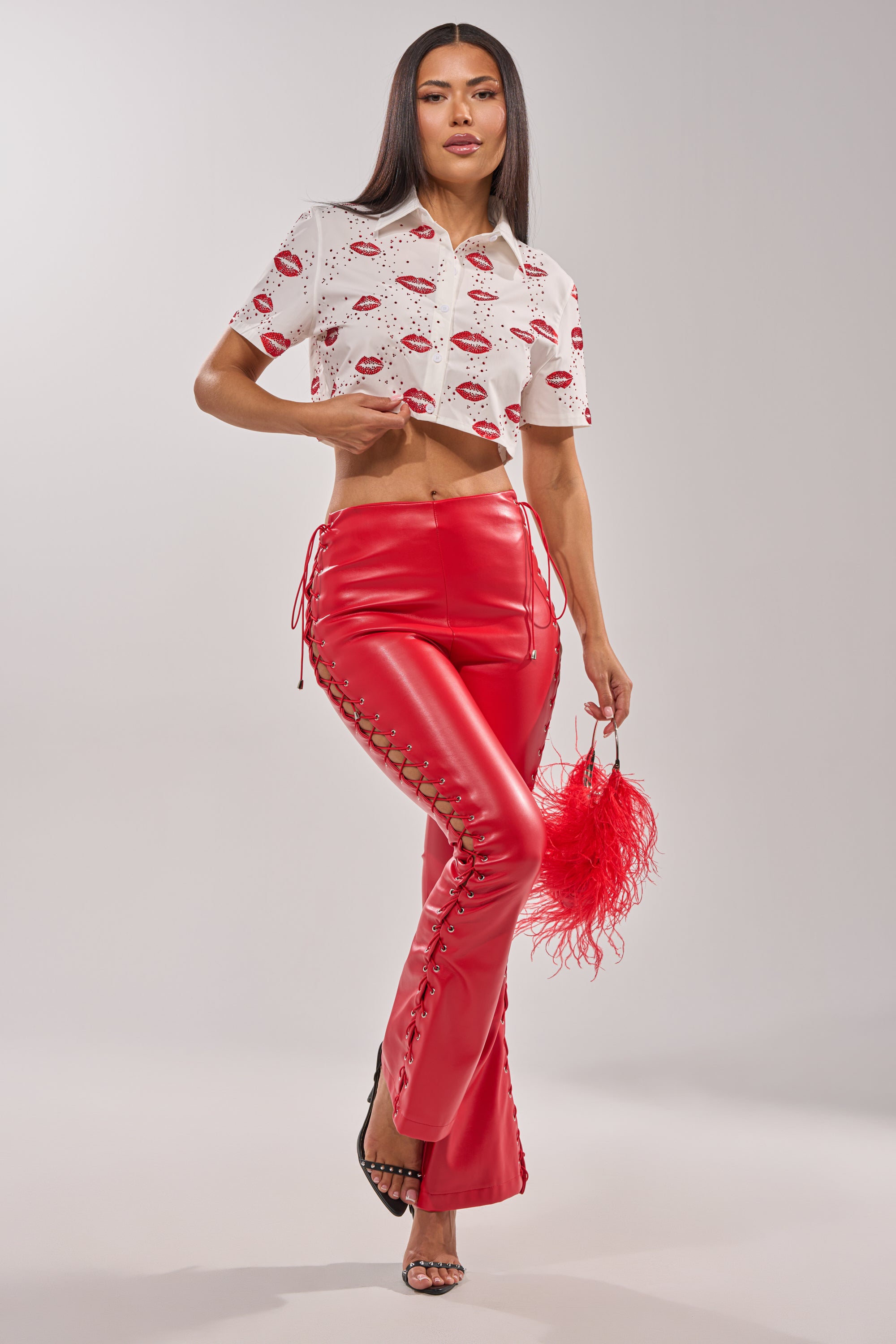 A woman poses in the KISS KISS RHINESTONE CROPPED BUTTON DOWN, bold red lace-up leather pants, a red feathered clutch, and black heeled sandals against a plain background. 10