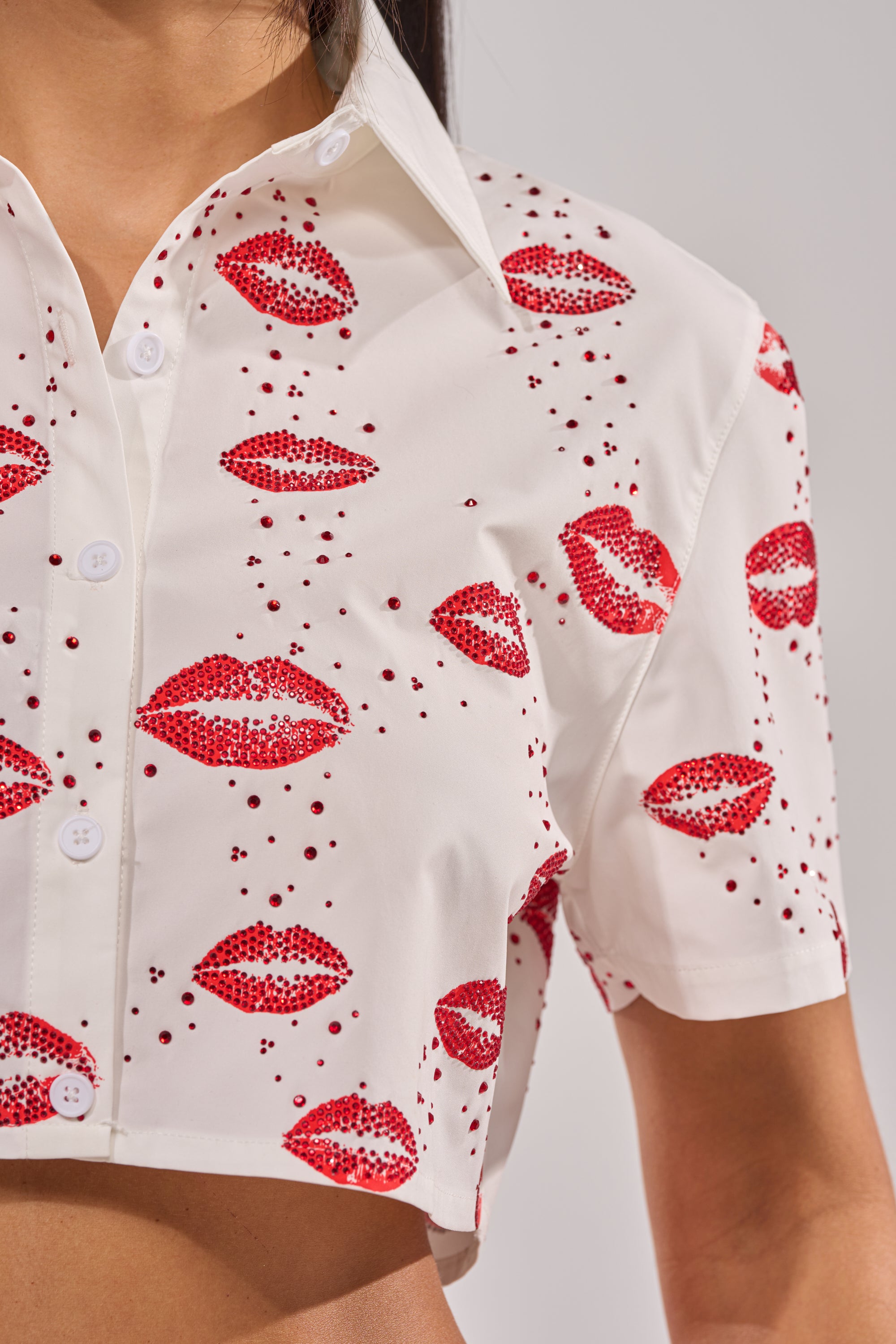 A close-up of a person wearing the KISS KISS RHINESTONE CROPPED BUTTON DOWN, showcasing its white fabric, playful red lips and dot pattern, sparkling rhinestones, and part of the torso. 11