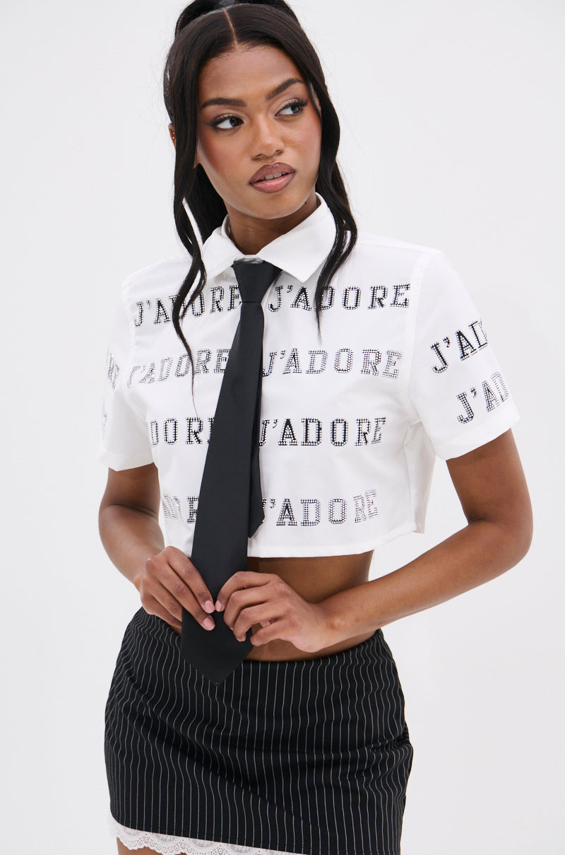 A person with long hair wears a J'ADORE RHINESTONE CROPPED BUTTON DOWN, a black tie, and a pinstripe skirt. French-inspired flair is conveyed as they look to the side against a plain background.