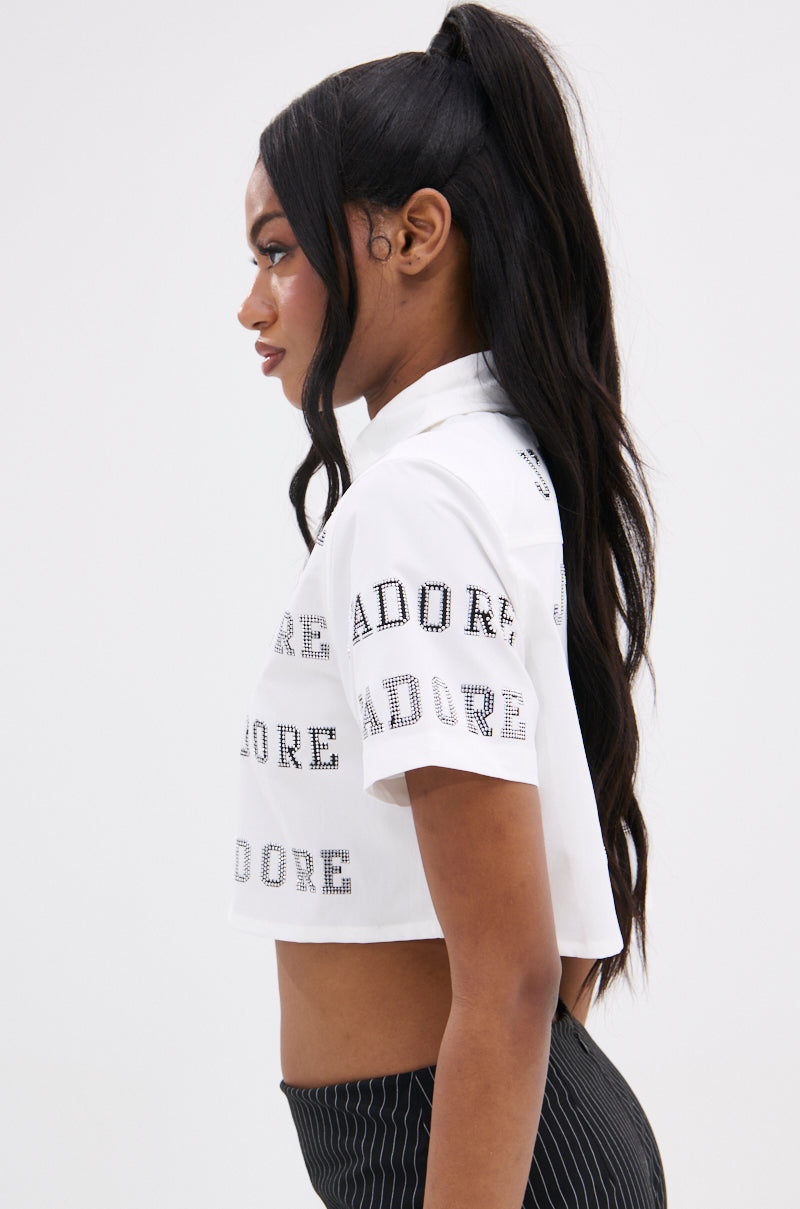 A person with long dark hair in a high ponytail wears the J'ADORE RHINESTONE CROPPED BUTTON DOWN against a plain backdrop, exuding a subtle French-inspired flair.