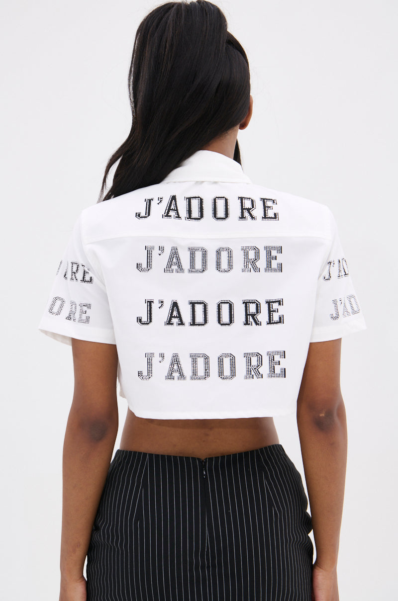 A person with long hair faces away, wearing the J'ADORE RHINESTONE CROPPED BUTTON DOWN featuring "J'ADORE" in black across the back and sleeves, showcasing French-inspired flair. Paired with black pinstripe pants against a plain light background.