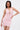 A woman with straight dark hair and tattoos poses confidently against a white background in the ALL EYES ON ME MINI DRESS IN PINK, featuring a ruched design and deep V neckline.