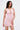 A woman wearing the ALL EYES ON ME MINI DRESS IN PINK, featuring a light pink hue, ruched design, and deep V neckline, stands against a white background, holding a small pink handbag at her side.