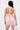 A woman with long dark hair models the ALL EYES ON ME MINI DRESS IN PINK, featuring floral lace bodycon details and a halter-neck cut. She faces away, holding a matching pink purse against a plain white background.