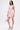 A woman stands against a white background, wearing the ALL EYES ON ME MINI DRESS IN PINK with ruched details and a deep V-neck. She pairs it with a shiny pink handbag and strappy pink heels with ruffle accents.