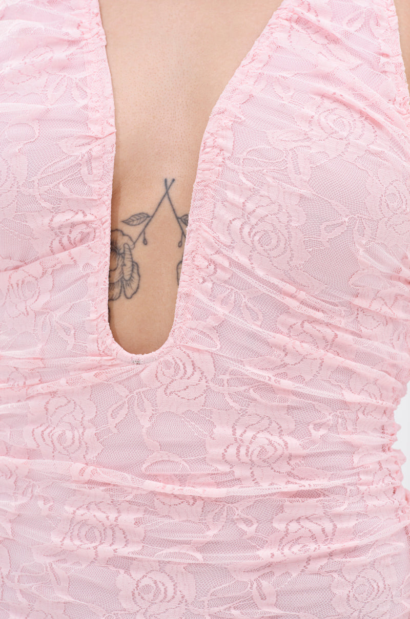 A close-up of a person wearing the ALL EYES ON ME MINI DRESS IN PINK, a light pink floral lace bodycon mini dress with a deep V neckline, revealing part of a floral tattoo on their chest.