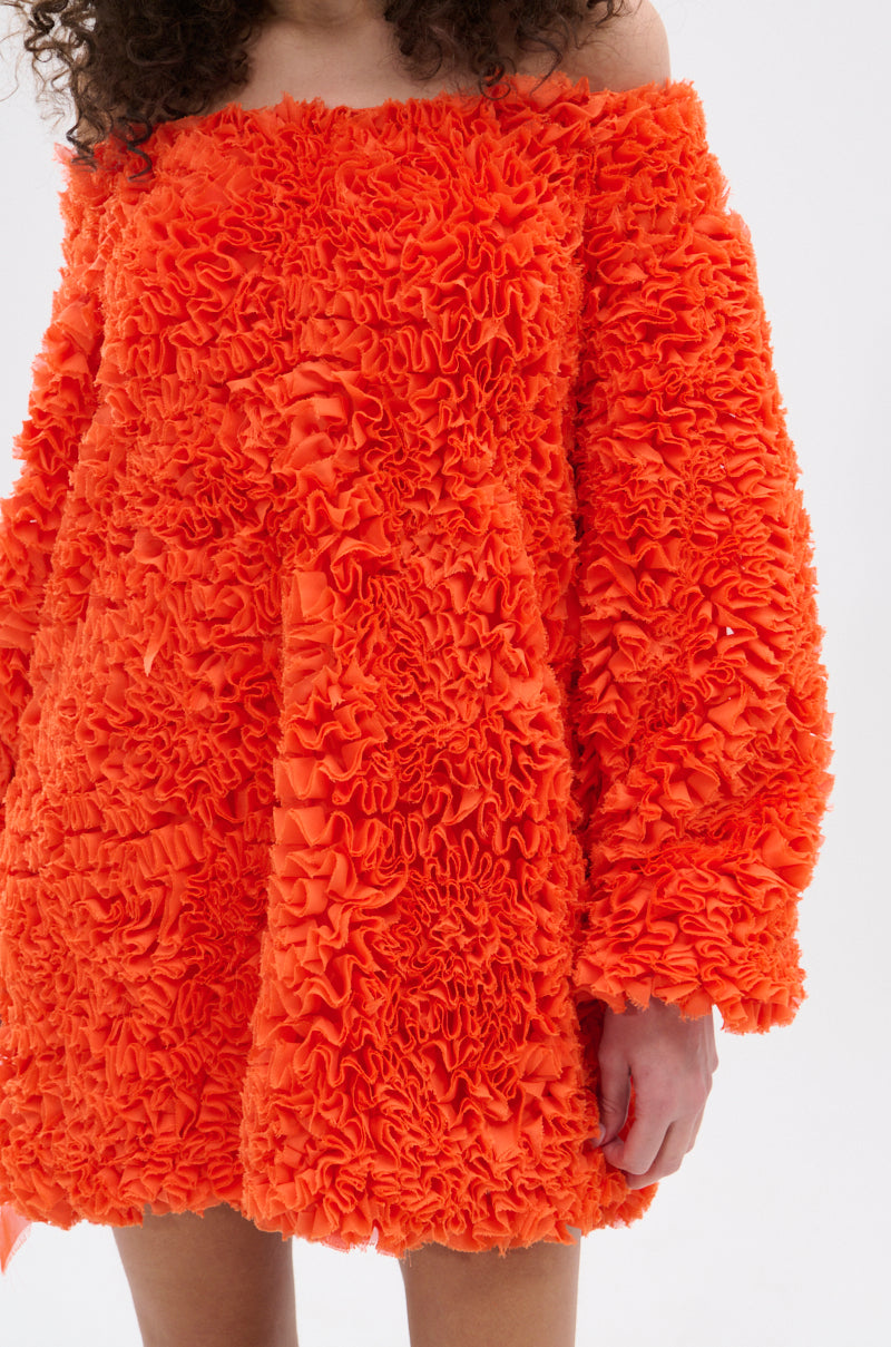 A person wears the IN THE NAME OF FASHION RUFFLE MINI DRESS, featuring bright orange textured ruffled fabric and off-the-shoulder long sleeves. Their face isn't visible, but the vibrant style commands attention.