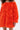 A person wears the IN THE NAME OF FASHION RUFFLE MINI DRESS, featuring bright orange textured ruffled fabric and off-the-shoulder long sleeves. Their face isn't visible, but the vibrant style commands attention.