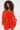 A woman with curly hair stands against a light background wearing the IN THE NAME OF FASHION RUFFLE MINI DRESS—an eye-catching orange, off-the-shoulder mini dress with long sleeves and textured fabric.