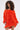 Someone with curly hair stands out in the IN THE NAME OF FASHION RUFFLE MINI DRESS, an off-the-shoulder, bright orange mini dress with textured fabric and long sleeves, set against a plain light background.