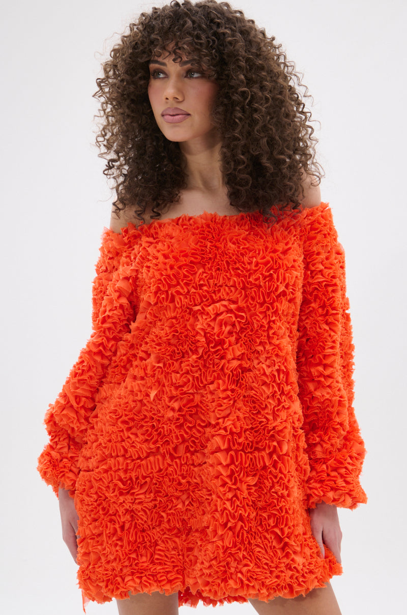 A woman with curly hair wears the IN THE NAME OF FASHION RUFFLE MINI DRESS—a bright orange, off-the-shoulder mini dress with textured, ruffled fabric—standing against a plain white background.