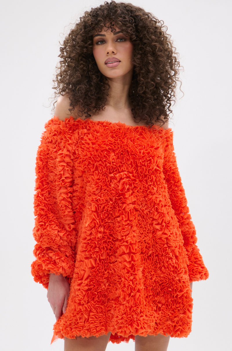 A woman with curly hair models the IN THE NAME OF FASHION RUFFLE MINI DRESS, an off-the-shoulder, bright orange mini dress with ruffled, fluffy fabric and long sleeves, posing against a plain light background.