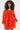 A woman with curly hair stands against a white background wearing the IN THE NAME OF FASHION RUFFLE MINI DRESS, a bright orange off-the-shoulder mini dress with textured ruffled fabric and long sleeves.