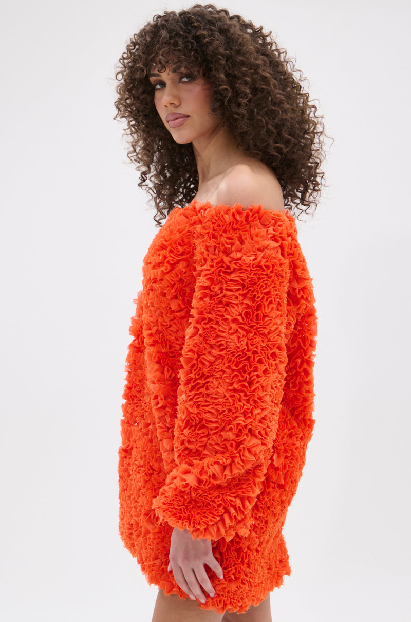 A woman with curly hair showcases the IN THE NAME OF FASHION RUFFLE MINI DRESS, a bright orange, off-the-shoulder mini with textured fabric and ruffled details, set against a plain white background.