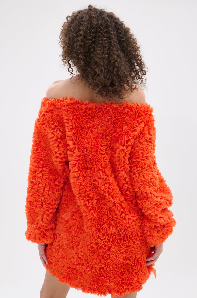 A person with curly hair is seen from behind, wearing the IN THE NAME OF FASHION RUFFLE MINI DRESS in bright orange with textured fabric and an off-the-shoulder design, standing out against a plain, light background.