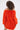 A person with curly hair is seen from behind, wearing the IN THE NAME OF FASHION RUFFLE MINI DRESS in bright orange with textured fabric and an off-the-shoulder design, standing out against a plain, light background.