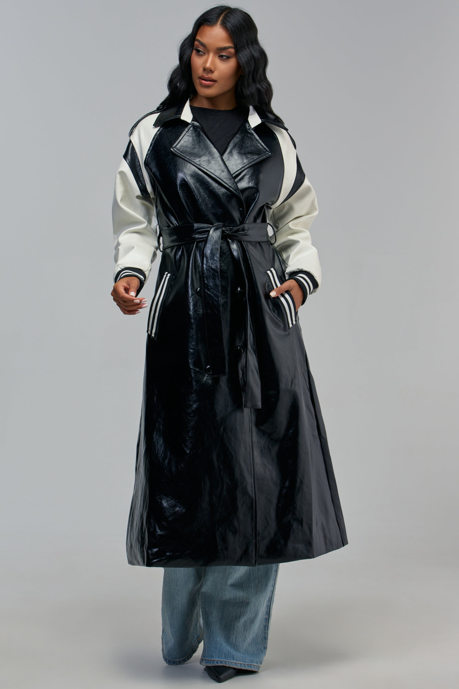 The CENTRAL PARK VARSITY TRENCH is a faux leather color block trench coat featuring a black body with white varsity-style sleeves, striped ribbed cuffs, and a belted waist.