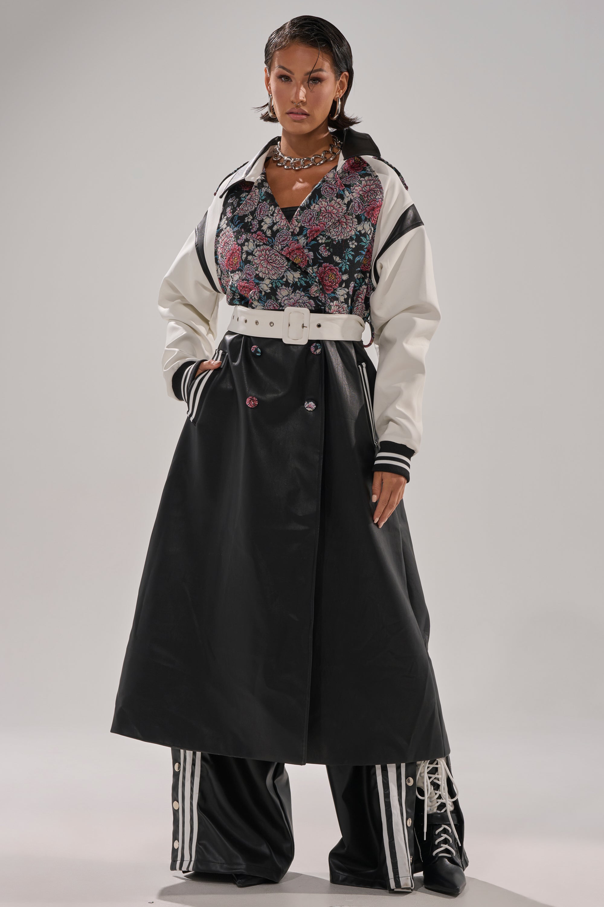A person stands confidently wearing the END GAME MIXED MEDIA TRENCH—a white belted jacket with black accents—over a floral top, long black skirt, and wide-legged striped pants. They style it with wet-look hair and a chunky necklace.