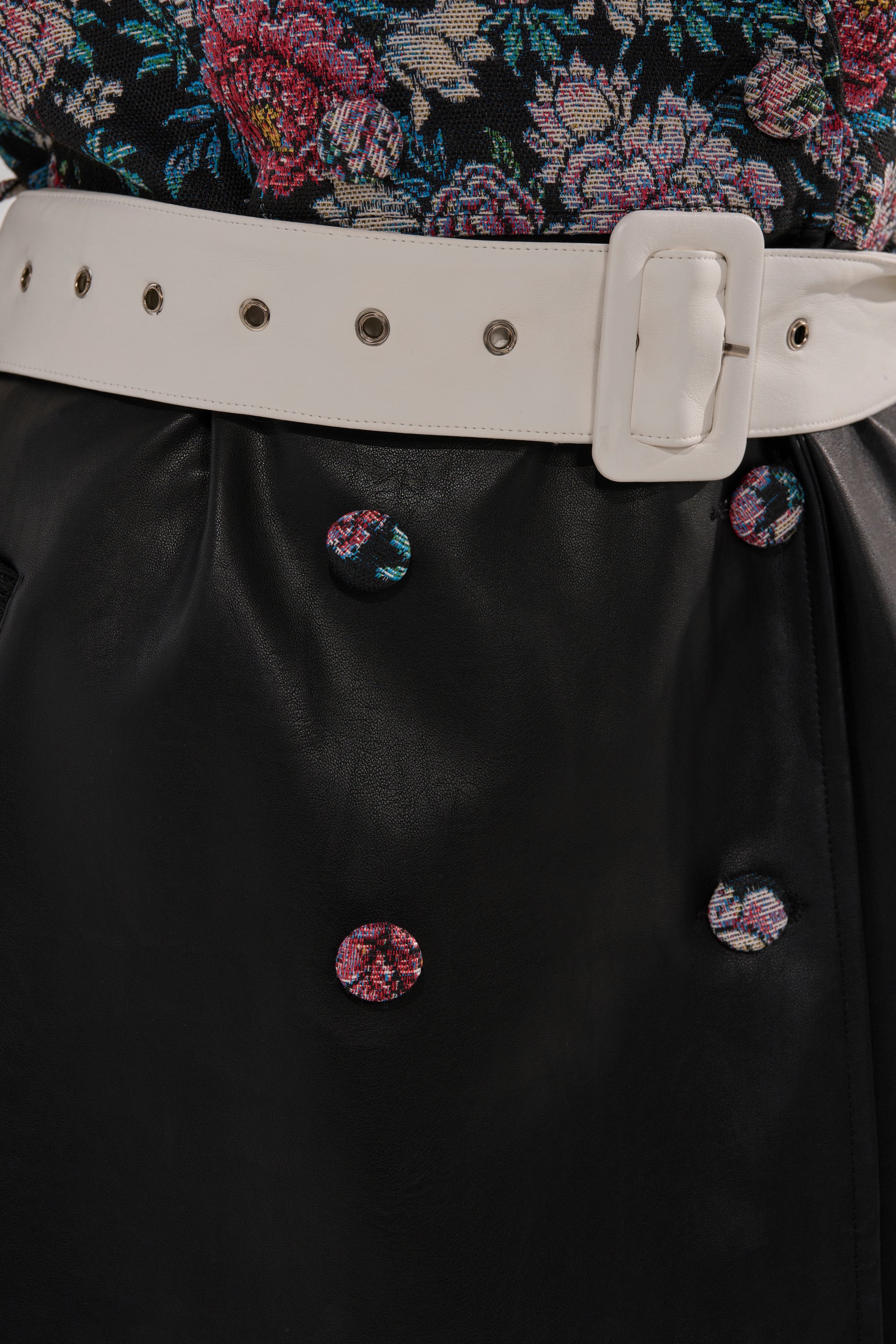 Close-up of the END GAME MIXED MEDIA TRENCH featuring black leather, floral fabric-covered buttons, a wide white belt with a square buckle, and a floral patterned top.