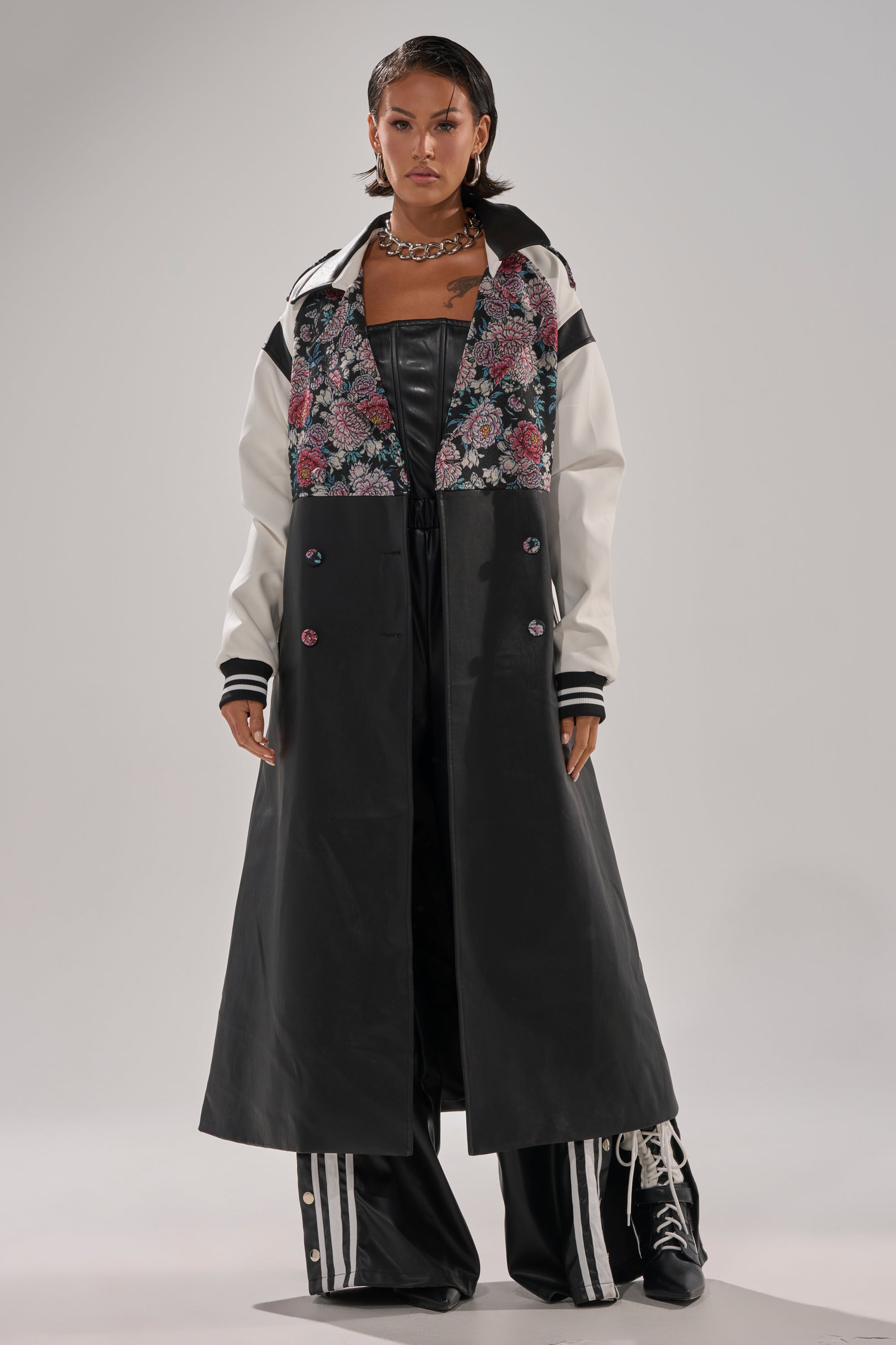 A person wears the END GAME MIXED MEDIA TRENCH, a long black and white coat with a floral-patterned upper section, paired with black leather pants, lace-up boots, and a chunky chain necklace, standing against a plain background.