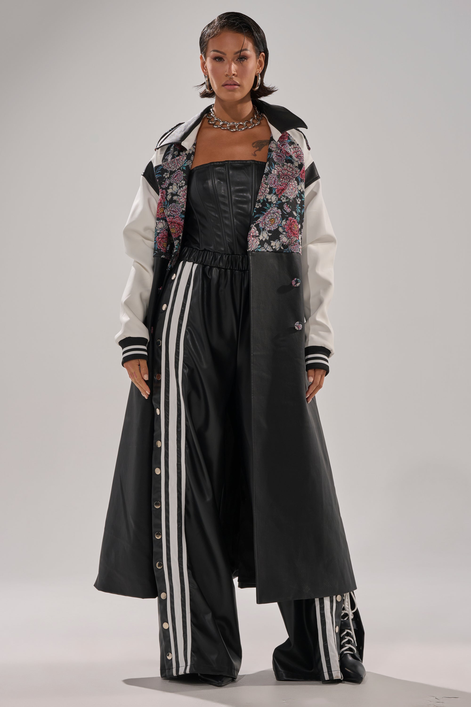 A person wears the END GAME MIXED MEDIA TRENCH—a long black and white floral coat—over a black strapless top and wide-leg pants with white side stripes, standing against a plain background.