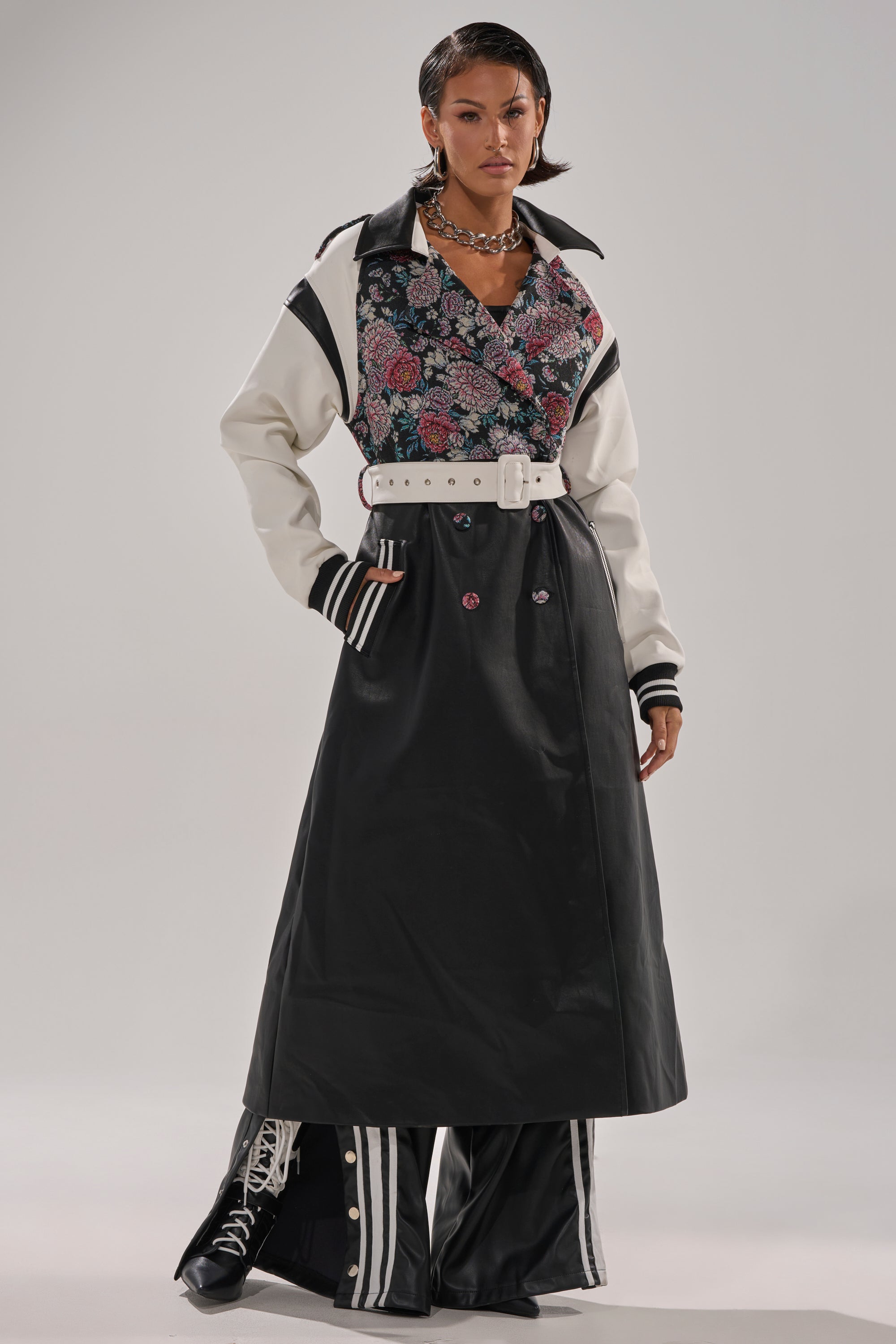 A woman stands confidently wearing the END GAME MIXED MEDIA TRENCH, a long black coat with white sleeves, paired with a floral shirt, wide white belt, striped wide-leg black pants, lace-up boots, and layered necklaces.