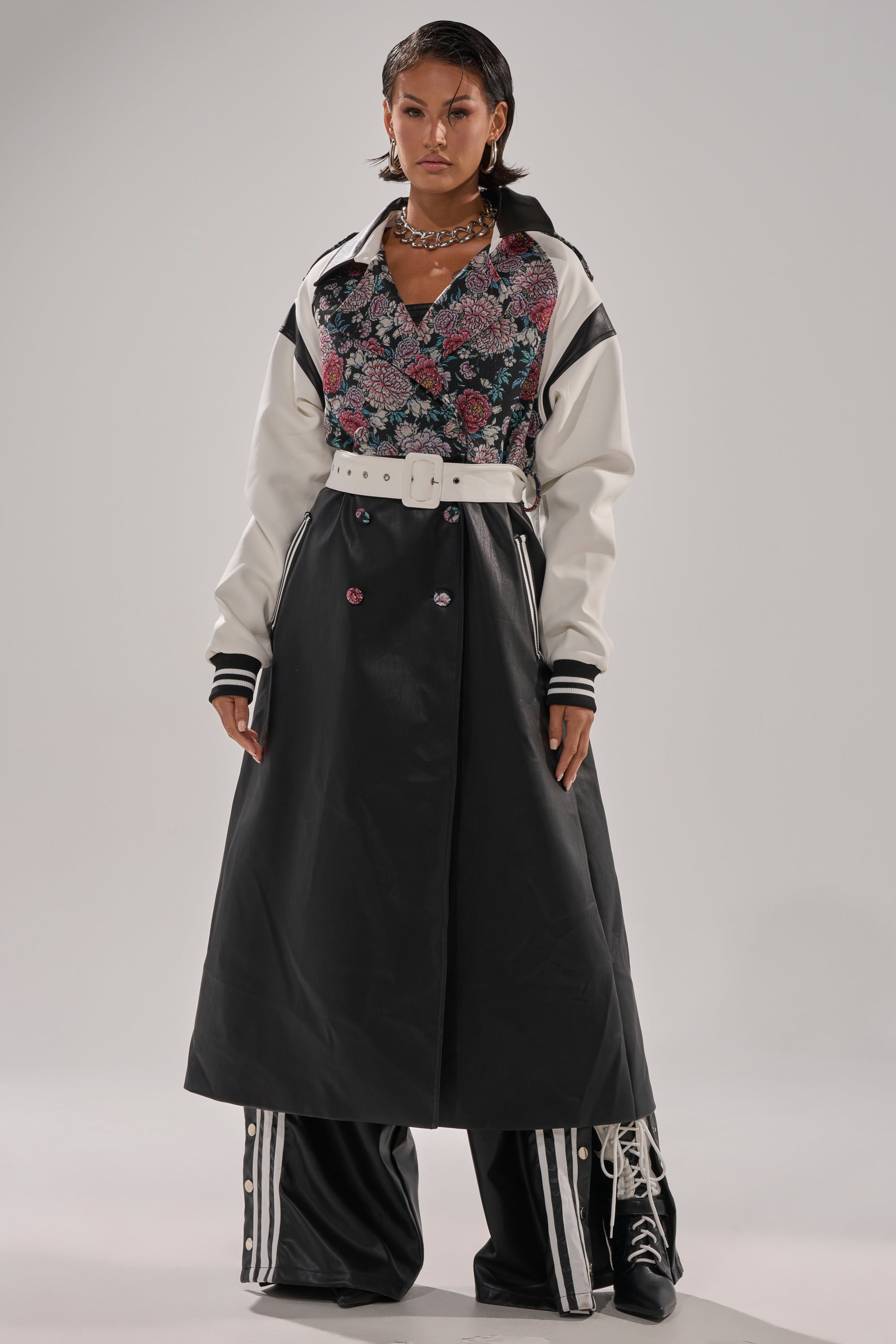 A person stands confidently wearing the END GAME MIXED MEDIA TRENCH, styled with a floral top, a long black buttoned skirt, wide black pants with white stripes underneath, and chunky black lace-up boots.