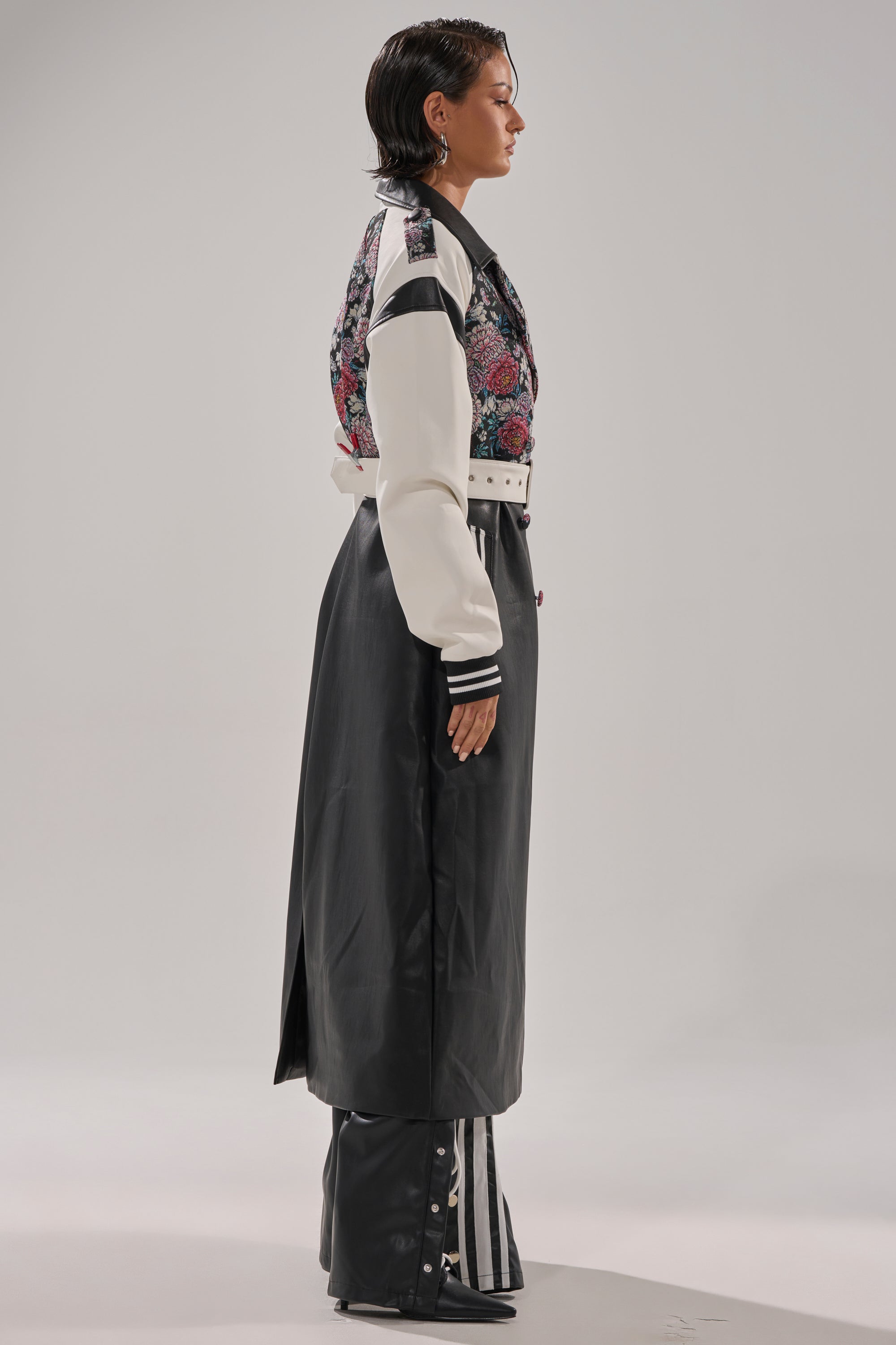 A person in profile wears the END GAME MIXED MEDIA TRENCH—a long black and white coat with floral patterns on top, a wide white belt, and matching black pants with white stripes and snap details—against a plain light background.